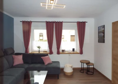 Gemütliches Wohnzimmer einer Ferienunterkunft in Bolsdorf (bei Hillesheim) mit dunkelgrauem Sofa und Fenster mit Vorhängen.