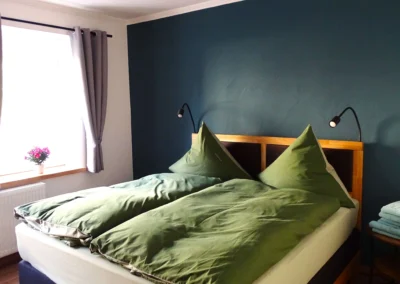 Helles Schlafzimmer einer Ferienunterkunft in Bolsdorf (bei Hillesheim) mit Doppelbett, grüner Bettwäsche, dunkelblauer Wand und Fenster mit Vorhängen.